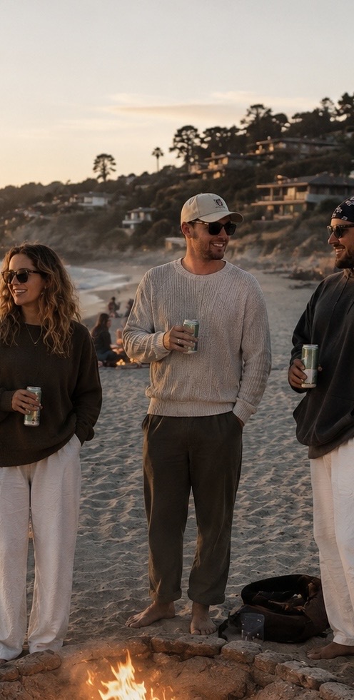 Friends in soft neutral layers gathered around a beach bonfire at sunset.