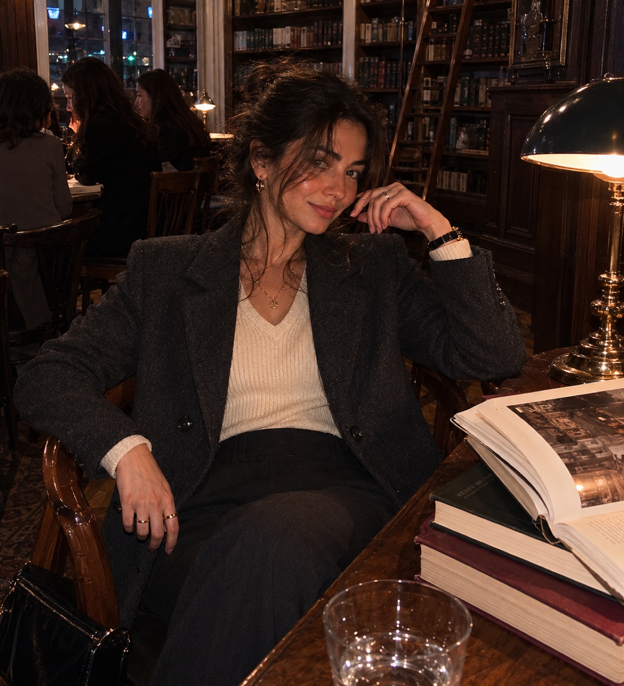 Woman in charcoal tailoring and a cream knit in a dark library lounge.